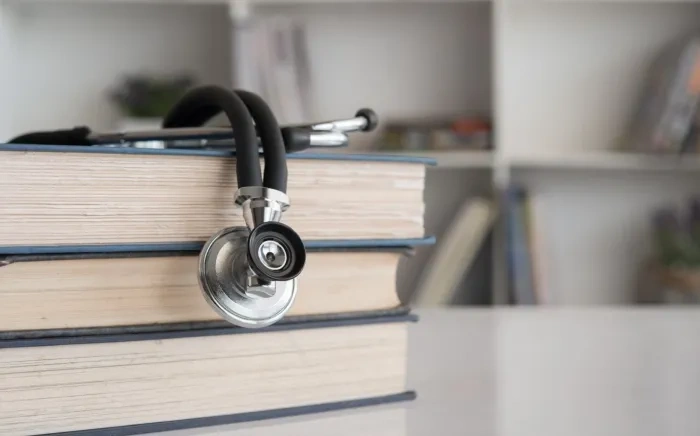 A stethoscope resting on a stack of books on a table, with a bookshelf in the background.