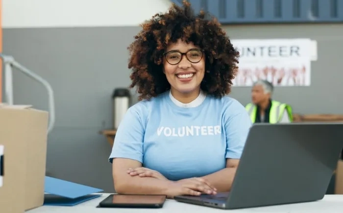 A smiling female volunteer wearing glasses and a blue T-shirt, sitting at a table with a laptop and tablet, with boxes in the background.