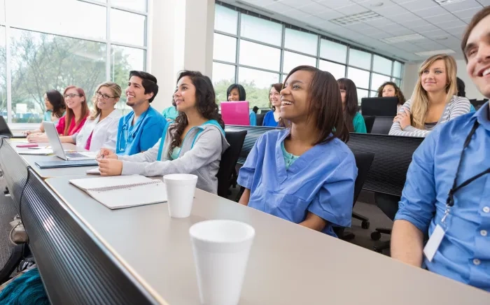 A diverse group of students or healthcare professionals sits in a classroom, smiling and attentively listening to a lecture, with notebooks and laptops on their desks.